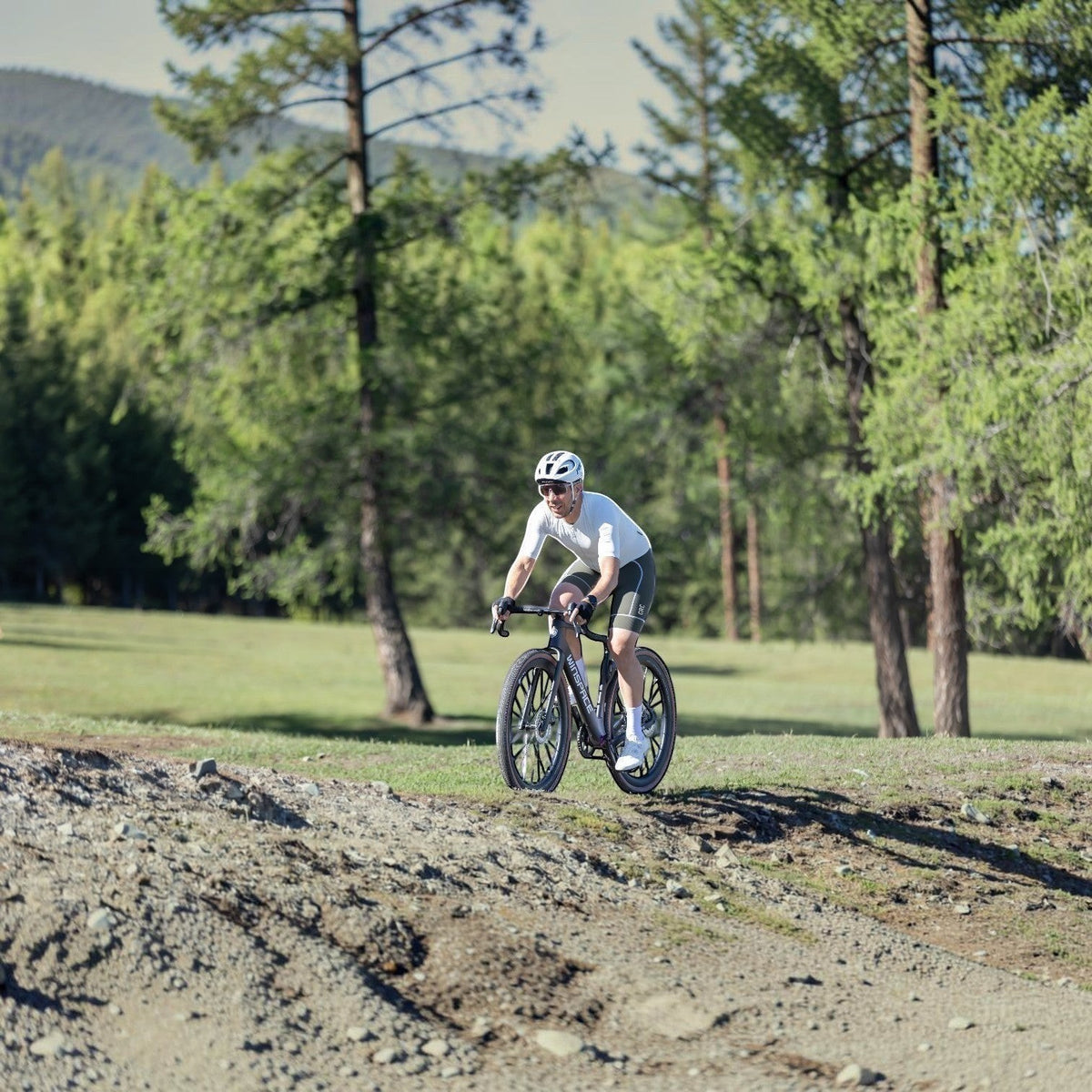 Cyclist riding a NorVelo G5 Gravel carbon bike on a dirt trail, wearing a white jersey and helmet in a forest setting.
