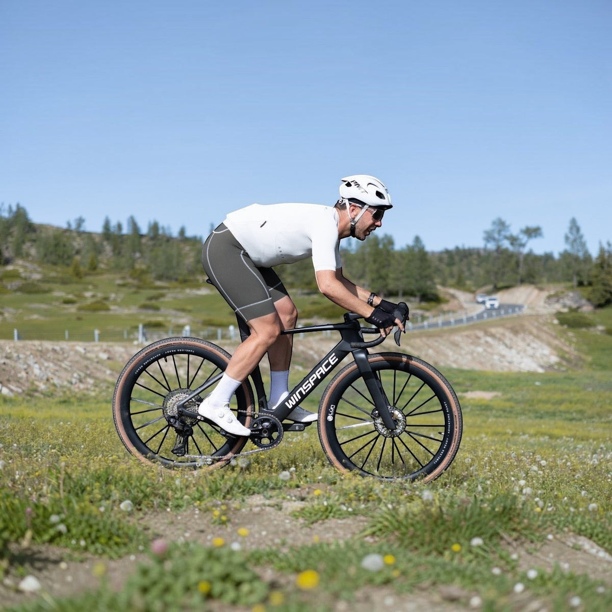 NorVelo G5 Gravel carbon bike ridden by a cyclist in white kit on a grassy trail with a winding road in the background.