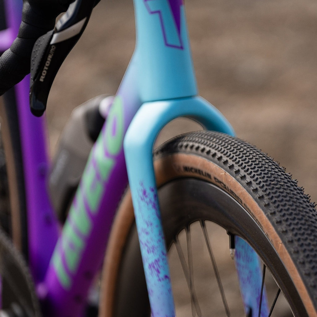 NorVelo Tavelo Grow aero gravel bike frame in blue and purple gradient with tan wall tires on a gravel path