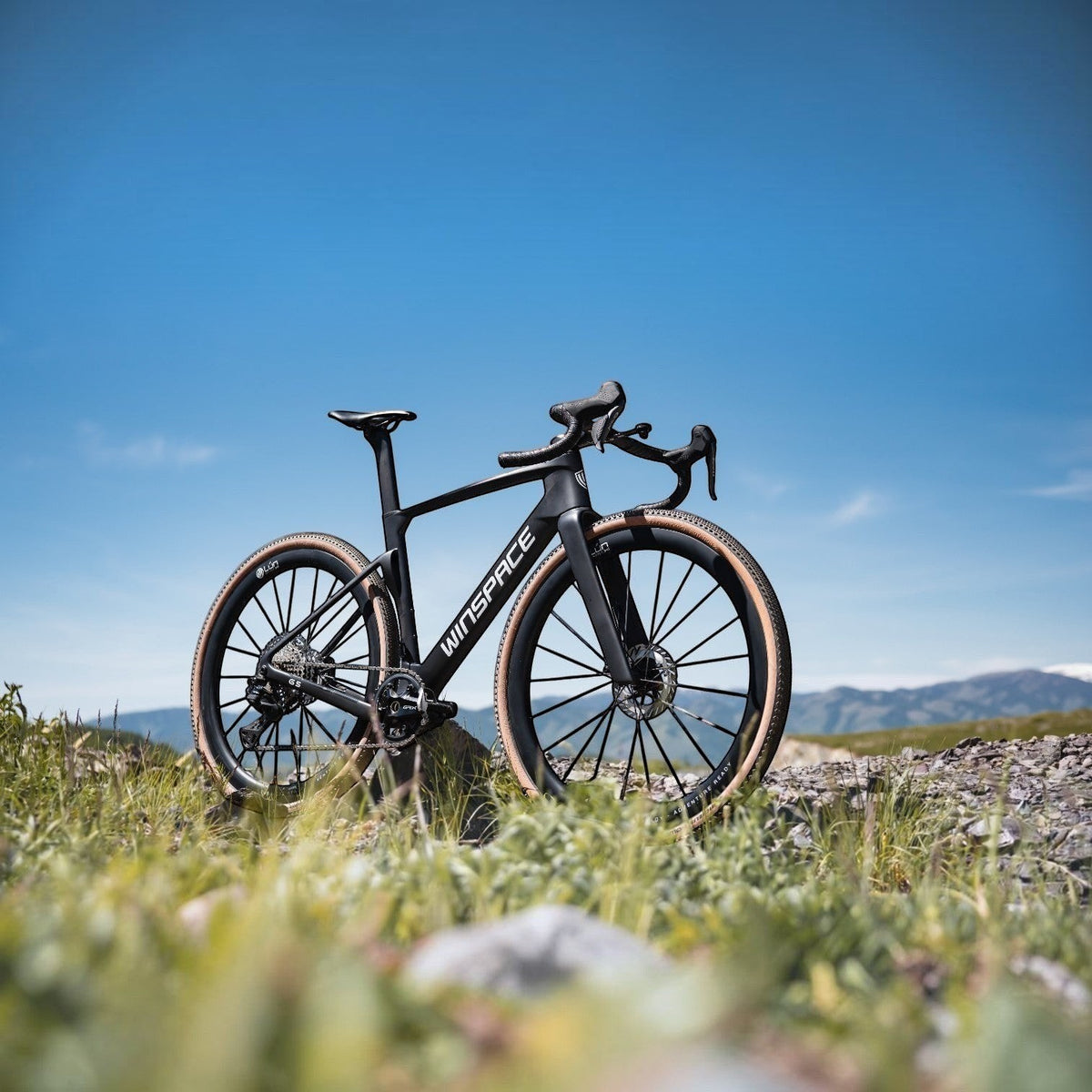 NorVelo G5 Gravel bike in matte black carbon with tan-wall tires, parked on a grassy hill under a clear blue sky.
