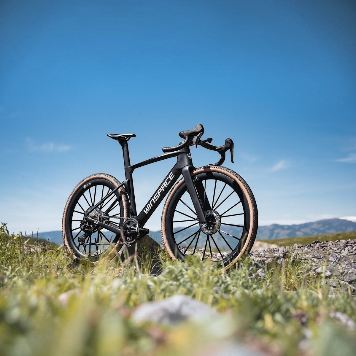 NorVelo G5 Gravel bike in matte black carbon with tan-wall tires, parked on a grassy hill under a clear blue sky.