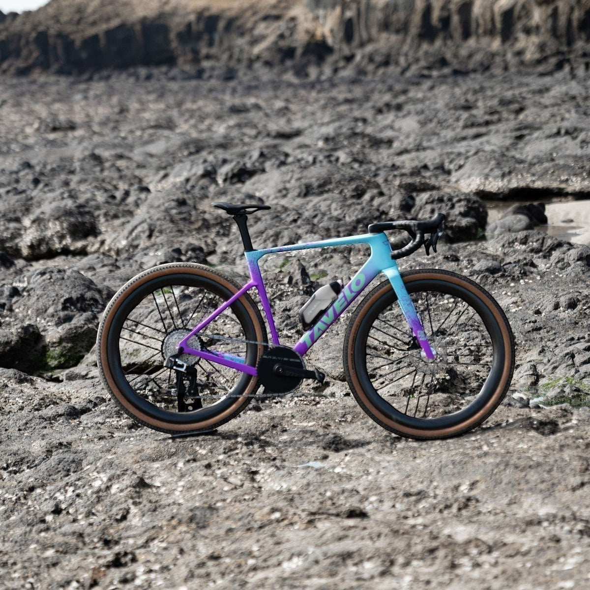NorVelo Tavelo Grow aero gravel bike with a purple and blue gradient carbon frame on a rocky terrain